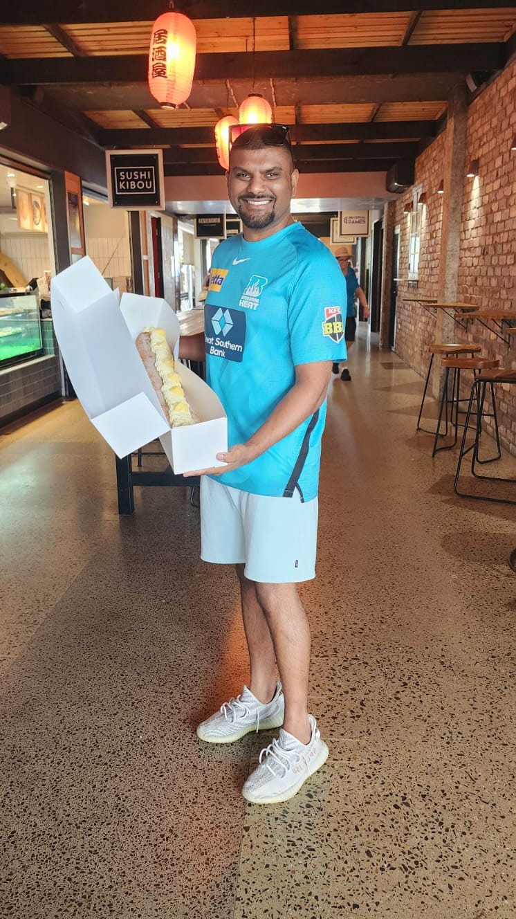 Zen standing in a walkway at The Wharf Mooloolaba while holding a white box displaying a long cream-topped donut pastry.