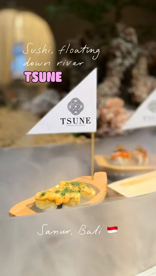 A wooden sushi boat with a Tsune paper flag floating along a misty water stream holding tamago sushi topped with mentaiko sauce.