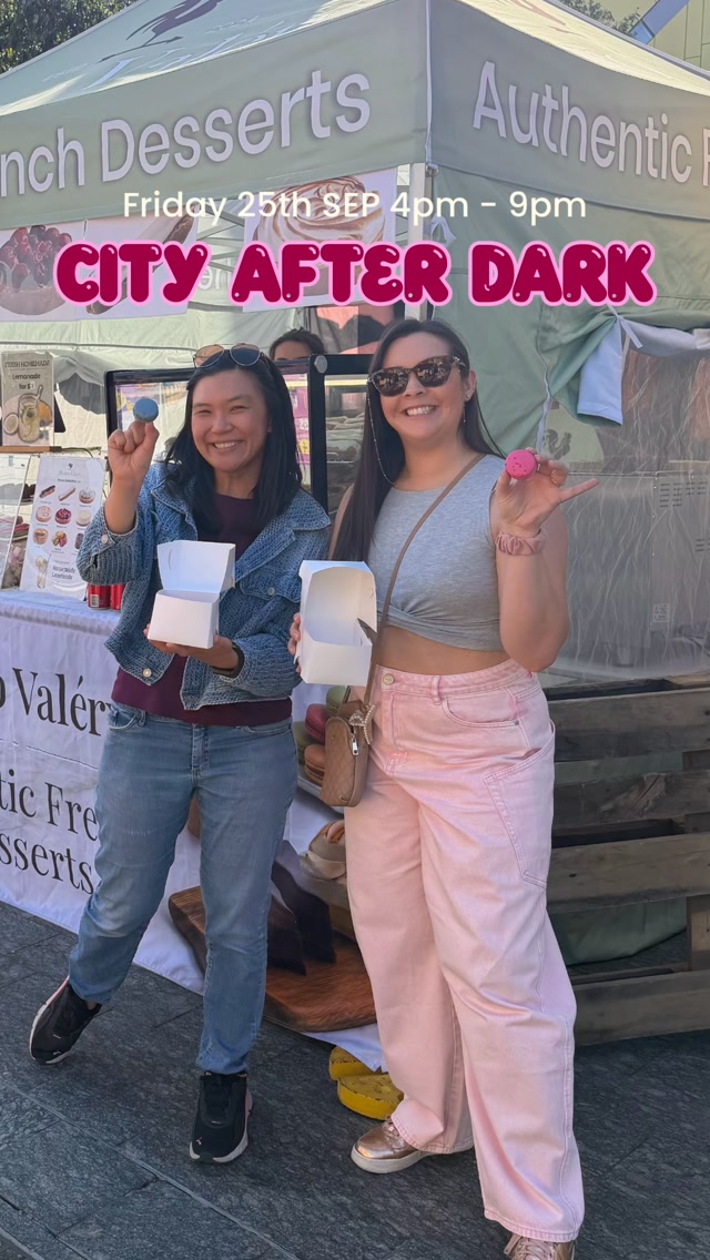 Two women smiling at a City After Dark market stall in front of a French Desserts sign, holding macarons and takeaway boxes.